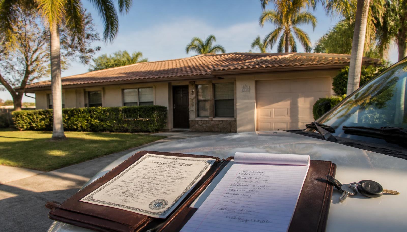 Property deed and handwritten loan figures on a leather folio resting on a truck hood in front of a paid-off concrete block Florida rental home with palm trees.