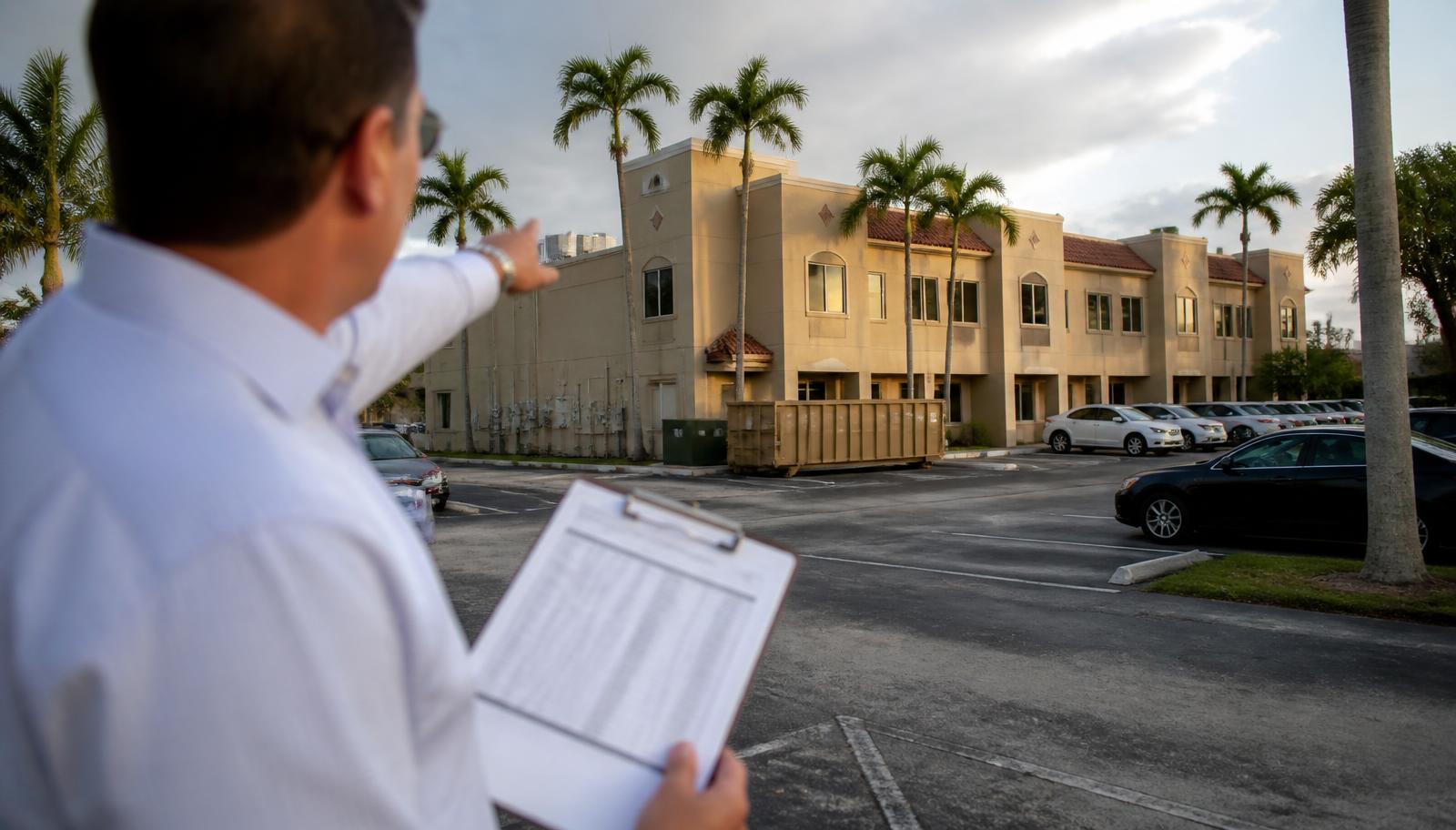 Commercial real estate broker holding a rent roll while looking at a two-story tan stucco multi-tenant Florida office and retail building with palm trees and a partly full parking lot.