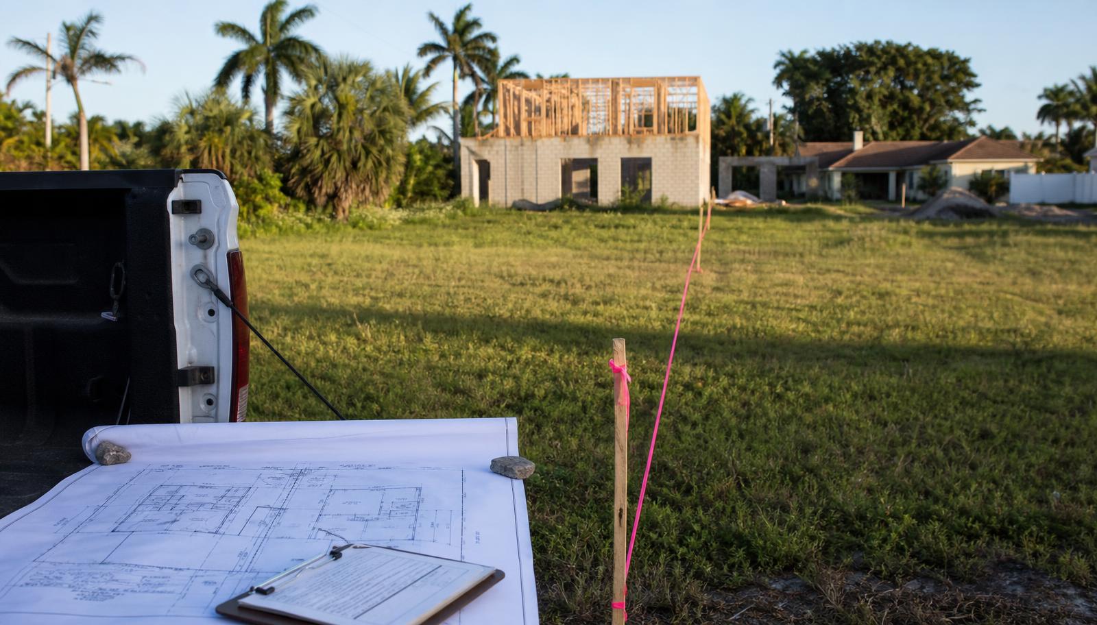 Lote baldío en Florida dividido con cinta rosada de agrimensor sobre estacas de madera, un levantamiento sobre la tapa de una camioneta y al fondo una casa en construcción con primer piso de bloque y segundo piso de madera.